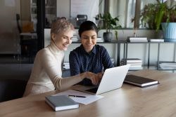 Two individuals sitting at a table in front of the laptop. One is mentoring the other.