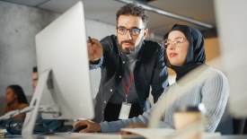 Two individuals sitting in front of a desktop computer. One is a professor and they are advising the student.